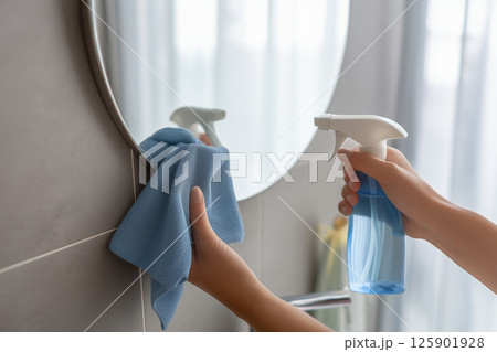 Person Cleaning a Bathroom Mirror with Spray Bottle and Blue Cloth in a Bright Modern Space 125901928