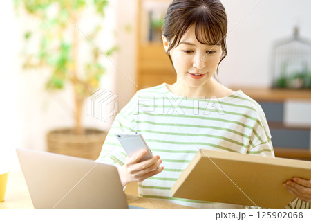 リビングでメール便の荷物を確認するスマホを持った若い女性 125902068