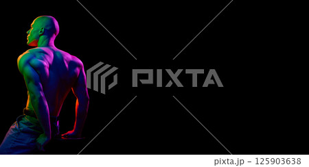 Side-angled view of young bald man, sting shirtless,demonstrating strong, muscular body against dark background in multi-colored neon light Side-angled view of young bald man, sting shirtless,demonstrating strong, muscular body against dark background in multi-colored neon light 125903638