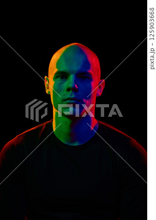 Portrait of young, handsome, bald man looking straight towards a camera with serious expression against black background in neon lights 125903668