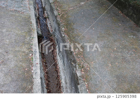 a narrow concrete drainage channel running through a paved area. The channel has a shallow flow of water and is flanked by worn concrete surfaces. The surrounding pavement is weathered with copyspace a narrow concrete drainage channel running through a paved area. The channel has a shallow flow of water and is flanked by worn concrete surfaces. The surrounding pavement is weathered with copyspace 125905398