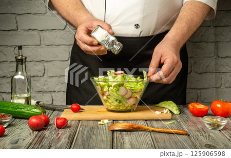 A chef mixes a vibrant salad using fresh ingredients like lettuce, radishes, cucumbers, and tomatoes, adding seasoning in a rustic kitchen environment 125906598