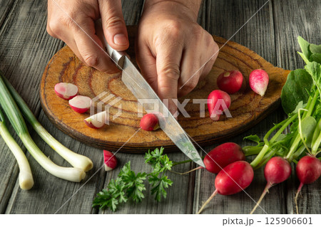 Hands skillfully slice vibrant radishes on a wooden board, surrounded by fresh greens and herbs, capturing the essence of healthy cooking in a warm kitchen atmosphere 125906601