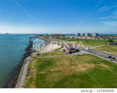 A coastal view showing a quiet seaside town with a rocky shoreline, green open fields, a road with passing vehicles, and residential buildings in the distance under a clear blue sky. 125906646