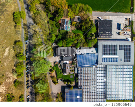 Aerial view of industrial buildings with solar panels, greenhouses, and residential houses surrounded by trees and fields, with a road and parked cars along the left side of the image. Aerial view of industrial buildings with solar panels, greenhouses, and residential houses surrounded by trees and fields, with a road and parked cars along the left side of the image. 125906649