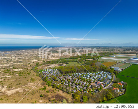 A stunning aerial view of a coastal town with red-tiled rooftops, neatly aligned houses, and a vast sandy beach stretching into the distance under a clear blue sky. A stunning aerial view of a coastal town with red-tiled rooftops, neatly aligned houses, and a vast sandy beach stretching into the distance under a clear blue sky. 125906652