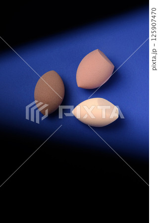 Three different cosmetic blenders on a blue background with a contrast shadow. Brown makeup sponges with pointed cutouts. Place for text. creative promotional photo for makeup artists. Three different cosmetic blenders on a blue background with a contrast shadow. Brown makeup sponges with pointed cutouts. Place for text. creative promotional photo for makeup artists. 125907470