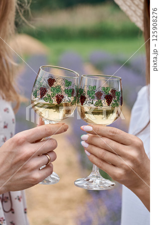 Cropped shot of two women cheering with glasses of white non-alcoholic homemade wine while standing in a lavender field. Glasses with a pattern. 125908276