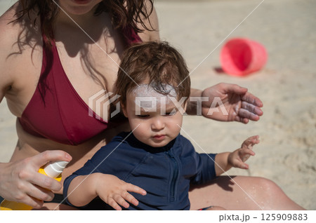 Caucasian woman applies sunscreen to her one-year-old son's skin.  125909883