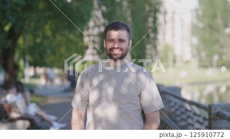 A cheerful young man stands outdoors in a sunny park, surrounded by green trees and a calm atmosphere. 125910772