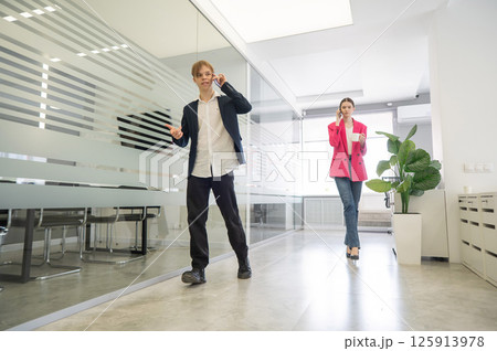 Two colleagues are walking through the office and talking on the phones. Two colleagues are walking through the office and talking on the phones. 125913978