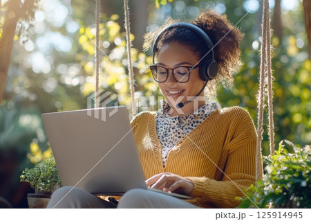 A woman wearing glasses and a headset is sitting on a swing and using a laptop 125914945