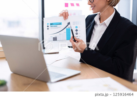 Business professional displaying economic data during meeting using printed chart, laptop on desk 125916464
