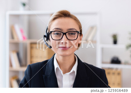 Smiling young caucasian woman wearing glasses and business suit with headset, standing in modern office setting providing customer service. Depicts professionalism, communication skills 125916489