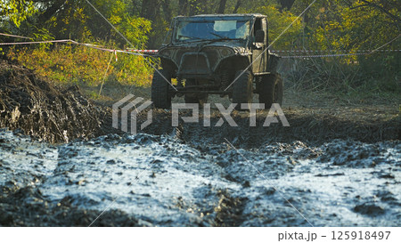 An off-road vehicle stopped in front of a large mud puddle during a race. The SUV is covered in mud and dust. The race takes place in the forest An off-road vehicle stopped in front of a large mud puddle during a race. The SUV is covered in mud and dust. The race takes place in the forest 125918497