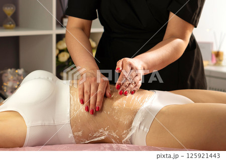Spa specialist massages client's wrapped abdomen with firm pressure. Spa specialist massages client's wrapped abdomen with firm pressure. 125921443
