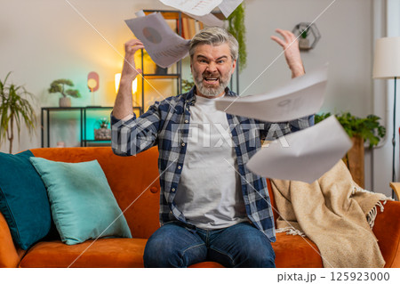 Dissatisfied senior man throwing out bills checks or papers documents while sitting on couch at home Dissatisfied senior man throwing out bills checks or papers documents while sitting on couch at home 125923000