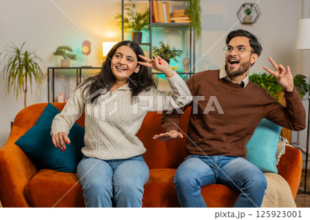 Smiling happy cheerful excited young Indian couple dancing enjoying weekend together on home couch 125923001