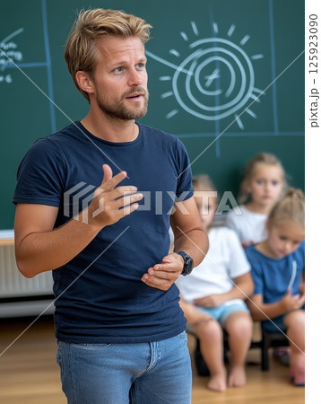 A young Caucasian male adult teacher gestures while speaking to a class of elementary-aged children 125923090