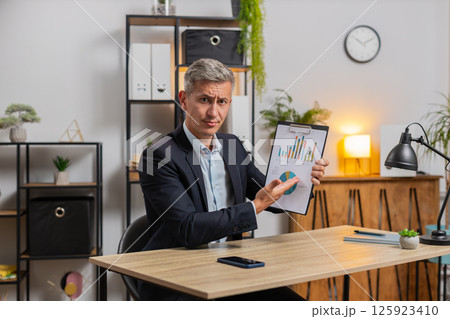 Confident young businessman guy showing graphical analytics during video call at home office desk Confident young businessman guy showing graphical analytics during video call at home office desk 125923410