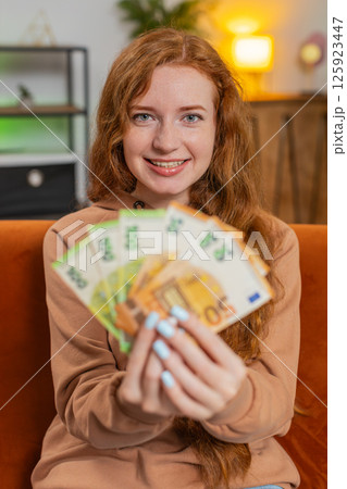 Successful rich young Caucasian woman holding euro bill money fan sitting on sofa couch at home Successful rich young Caucasian woman holding euro bill money fan sitting on sofa couch at home 125923447