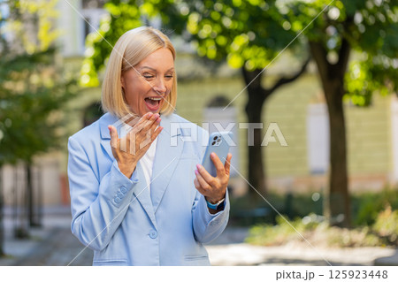 Caucasian mature businesswoman looking shocked while reading good news on smartphone on city street Caucasian mature businesswoman looking shocked while reading good news on smartphone on city street 125923448