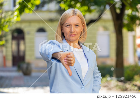 Upset mature Caucasian blonde businesswoman in formal suit looking camera and showing thumb down Upset mature Caucasian blonde businesswoman in formal suit looking camera and showing thumb down 125923450