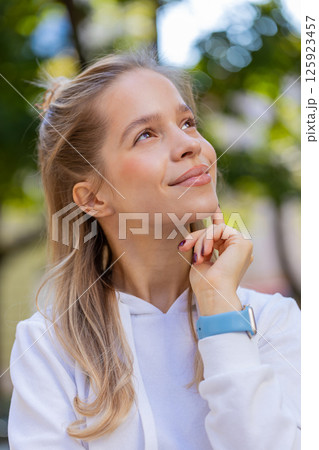 Thoughtful smiling young woman with pensive expression touching chin looking around on city street Thoughtful smiling young woman with pensive expression touching chin looking around on city street 125923457