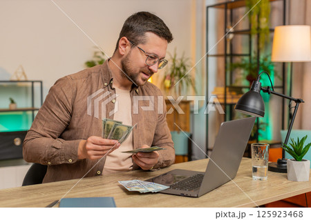 Rich happy Caucasian man counting money cash using laptop calculate domestic bills at home table Rich happy Caucasian man counting money cash using laptop calculate domestic bills at home table 125923468