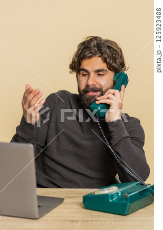 Happy businessman sitting at table talking on wired telephone and using laptop on beige background Happy businessman sitting at table talking on wired telephone and using laptop on beige background 125923488