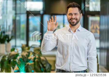 Happy young Caucasian businessman looking at camera and waves hand hello in modern office lobby Happy young Caucasian businessman looking at camera and waves hand hello in modern office lobby 125923536