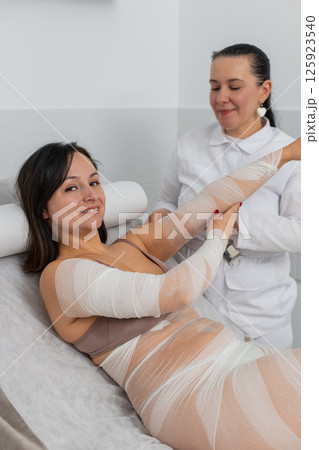 Cosmetologist warping woman hand with compression elastic bandage body mask regeneration weight loss Cosmetologist warping woman hand with compression elastic bandage body mask regeneration weight loss 125923540