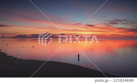 Breathtaking aerial view of a vibrant sunset reflected in calm ocean waters, with a lone man standing on the shoreline, creating a serene and picturesque scene in a tropical paradise 125924699