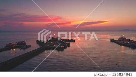 Ferry departing from the pier at sunset, casting vibrant hues of pink and orange across the sky. Calm sea and majestic mountains create a serene backdrop, enhancing the tranquil evening scene 125924942