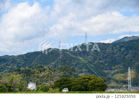A scenic landscape featuring lush green mountains under a partly cloudy sky. Tall electricity transmission towers line the ridges 125925050