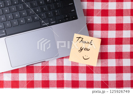 Remote workspace with laptop and Thank you note on red and white checkered table 125926749