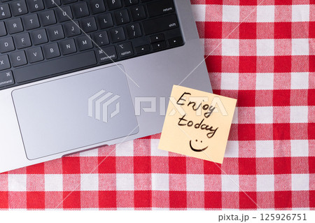 Sticky note with "Enjoy today" text next to laptop on checkered table for positive daily reminder 125926751