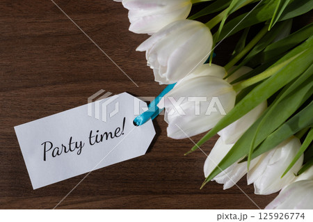 Tag with Party time text attached to white tulips on wooden background ideal for party invitations event announcements celebration messages or festive spring promotions Tag with Party time text attached to white tulips on wooden background ideal for party invitations event announcements celebration messages or festive spring promotions 125926774