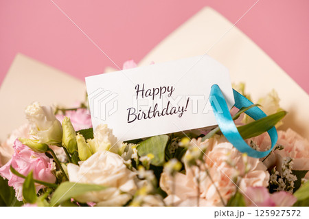 beautiful birthday bouquet with a handwritten "happy birthday" card attached by a blue ribbon, featuring delicate pastel flowers against a soft pink background 125927572