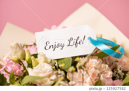 cheerful bouquet of pastel flowers with a handwritten enjoy life message card tied with a blue ribbon, radiating positivity and joy on a soft pink background 125927582