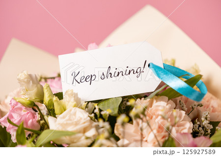 motivational message keep shining on a handwritten card, attached with a blue ribbon to a fresh floral bouquet of pastel roses and lisianthus against a soft pink background 125927589