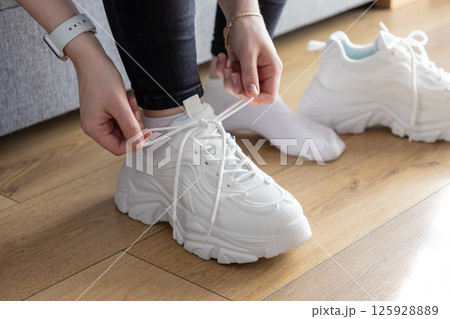Person's hands fastening white laces on chunky training sneakers indoors, indicating preparation for comfortable and supportive exercise Person's hands fastening white laces on chunky training sneakers indoors, indicating preparation for comfortable and supportive exercise 125928889