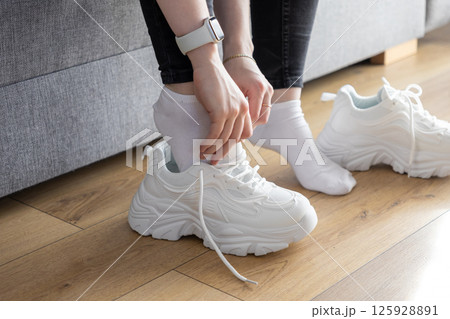 Close up of hands putting on white chunky athletic sneakers indoors, showing preparation for comfortable fitness activity 125928891