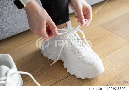 Close up of person's hands tying white laces on chunky training sneakers indoors, preparing for comfortable sport activity 125928895
