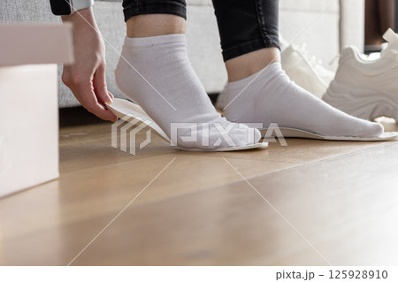 Close up of hands positioning white orthopedic insole onto socked foot indoors, enhancing comfort and support for sport shoes Close up of hands positioning white orthopedic insole onto socked foot indoors, enhancing comfort and support for sport shoes 125928910