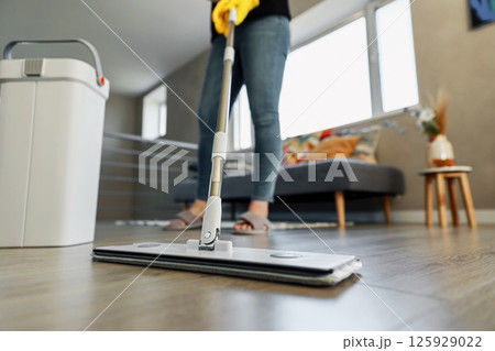 Woman mopping, wiping sleek hardwood floor in modern home setting. Girl wearing yellow gloves. Housewife uses professional cleaning supplies: mop, plastic bucket with wringer in cozy living house room 125929022