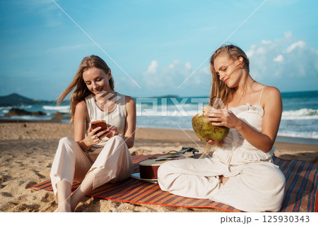 Young Women Smiling on the Beach with Drinks and Smartphones 125930343