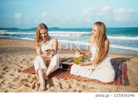Two Women Relaxing on the Beach with Refreshing Coconut  125930346