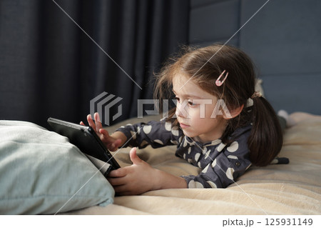 Focused little girl lies on bed and interacts with tablet screen, her face serious and intent 125931149