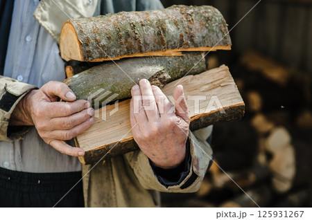 Rough, veined hands carry freshly chopped logs. Behind them, firewood is piled high. Rough, veined hands carry freshly chopped logs. Behind them, firewood is piled high. 125931267
